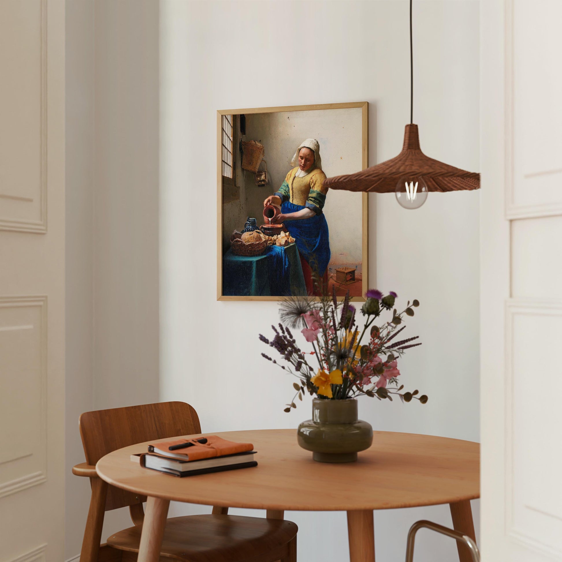 a cozy dining area with a wooden table, a vase of flowers, and a framed painting of a woman in a blue dress. The table is set with a book and a notebook, creating a comfortable and inviting atmosphere.