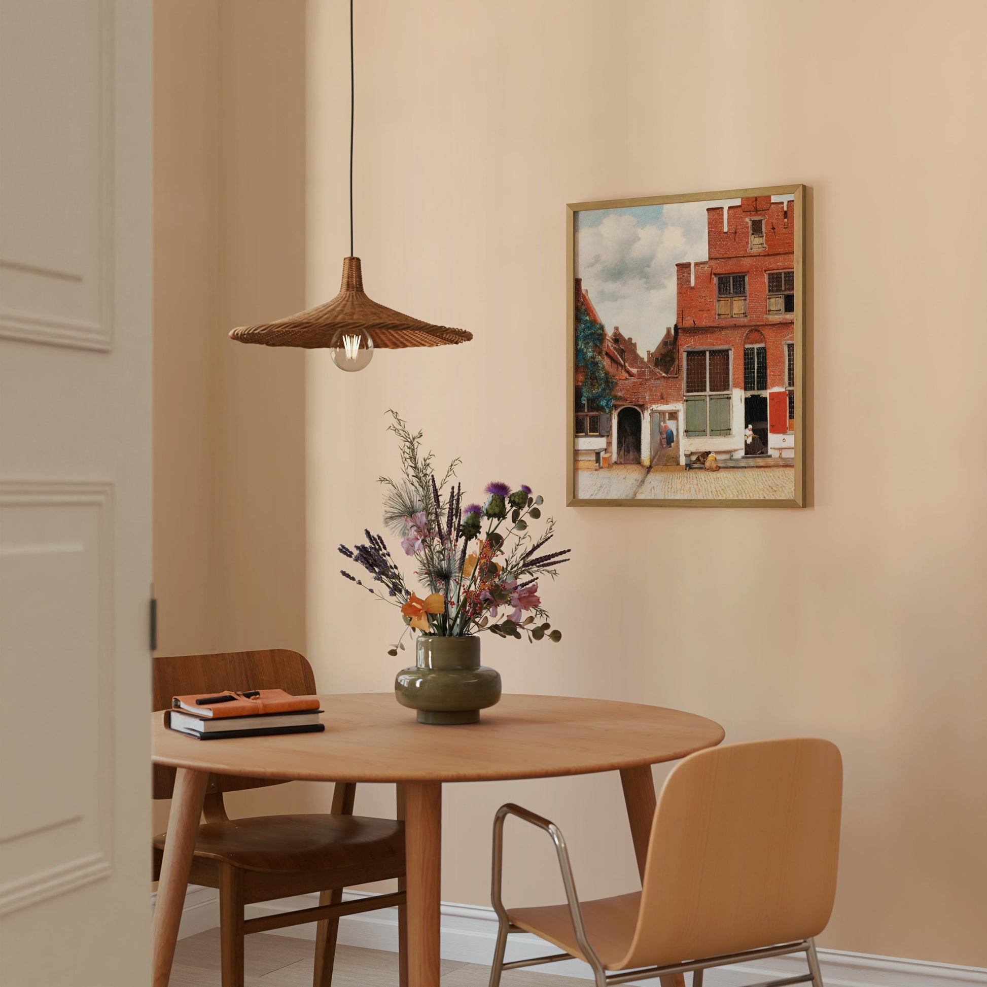 a cozy dining area with a wooden table, two chairs, and a vase of flowers. A painting of a building is hanging on the wall above the table, and a hanging light fixture is also present in the room.